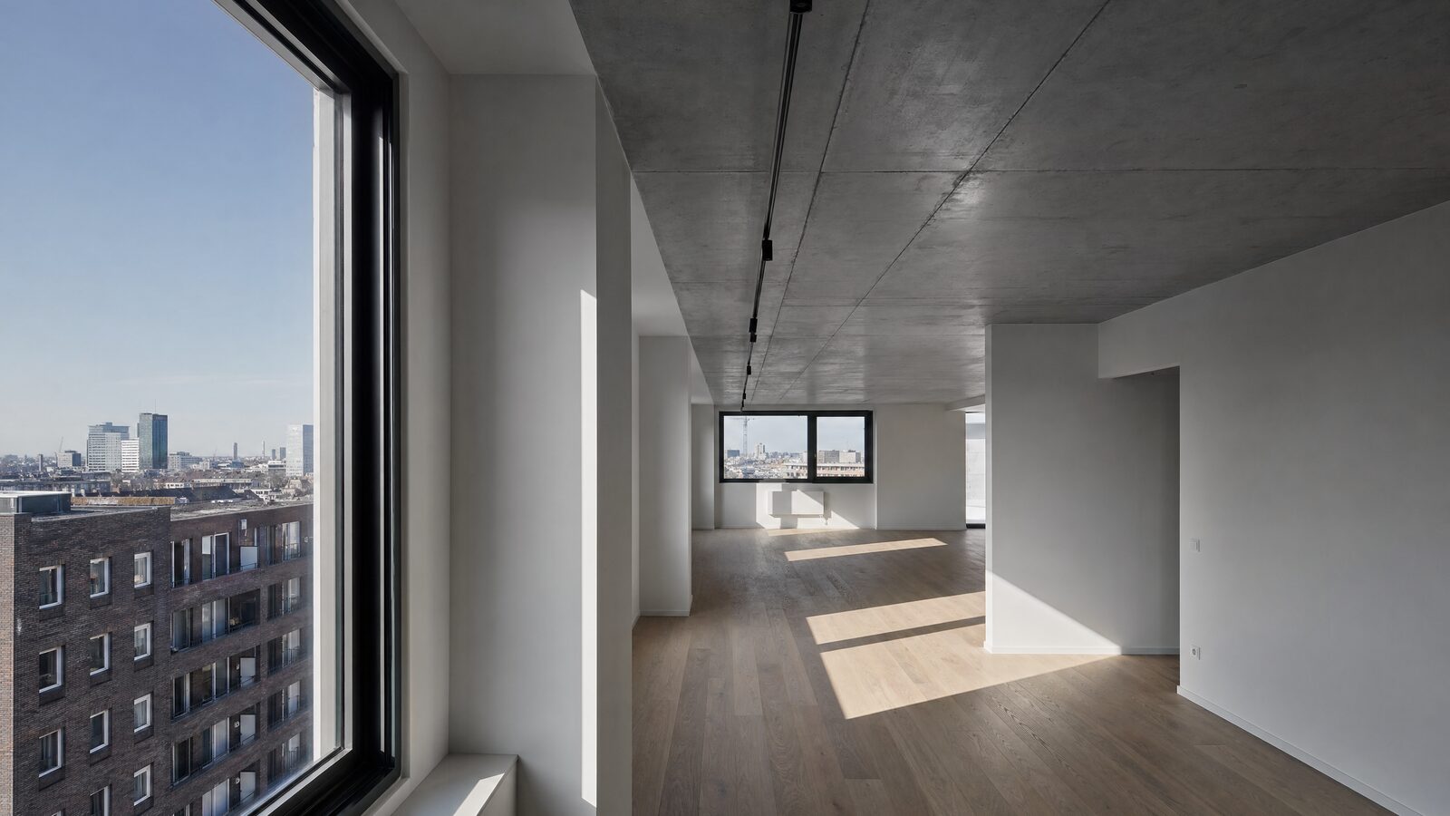 Gerenoveerde open leefruimte in een appartement met zichtbaar betonnen plafond, eiken vloer en een groot bestaand raam
