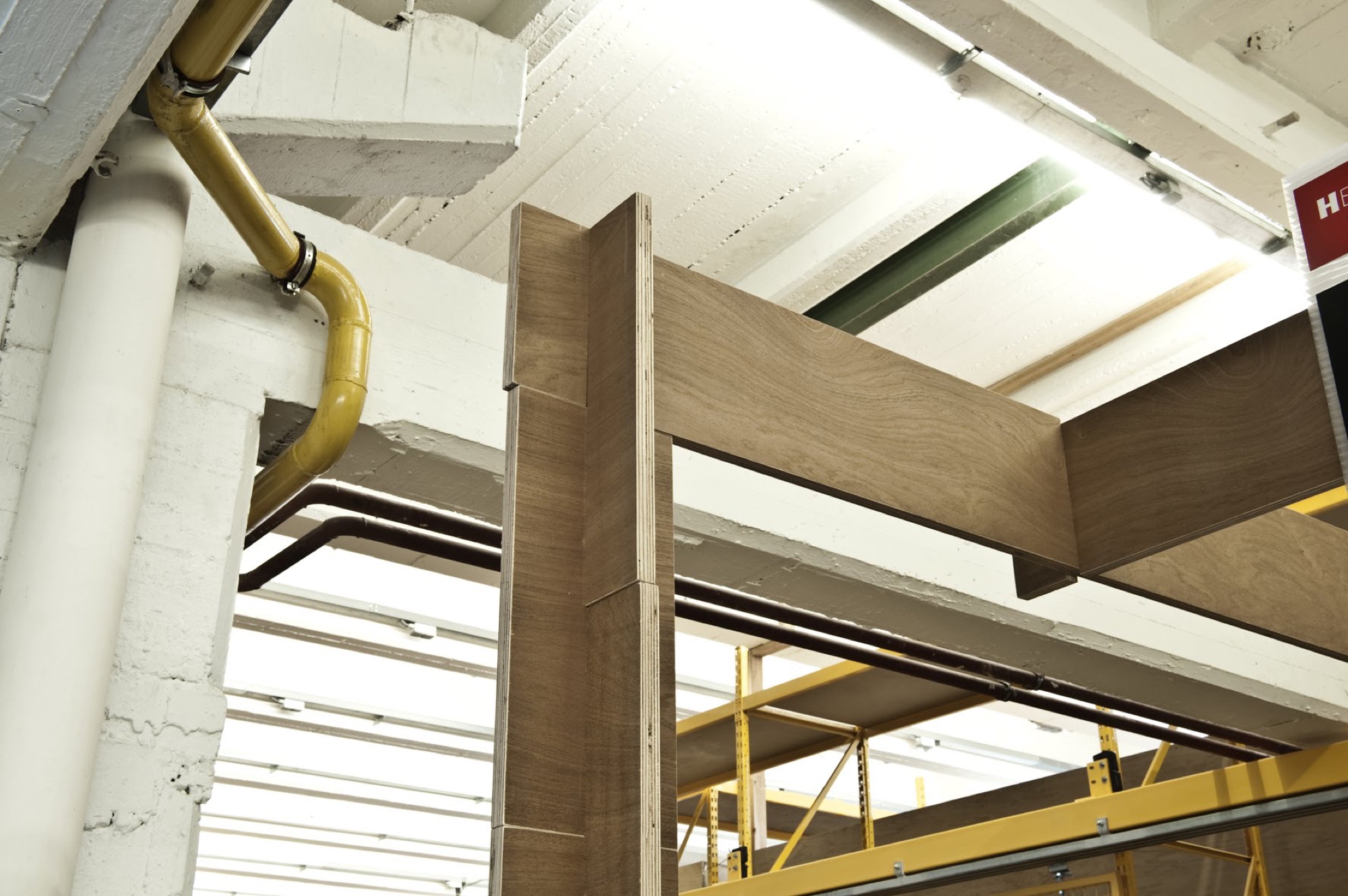 Detail van een houten portiek van BUDA.lab tegen de witte betonstructuur, met een gele metalen constructie op de achtergrond
