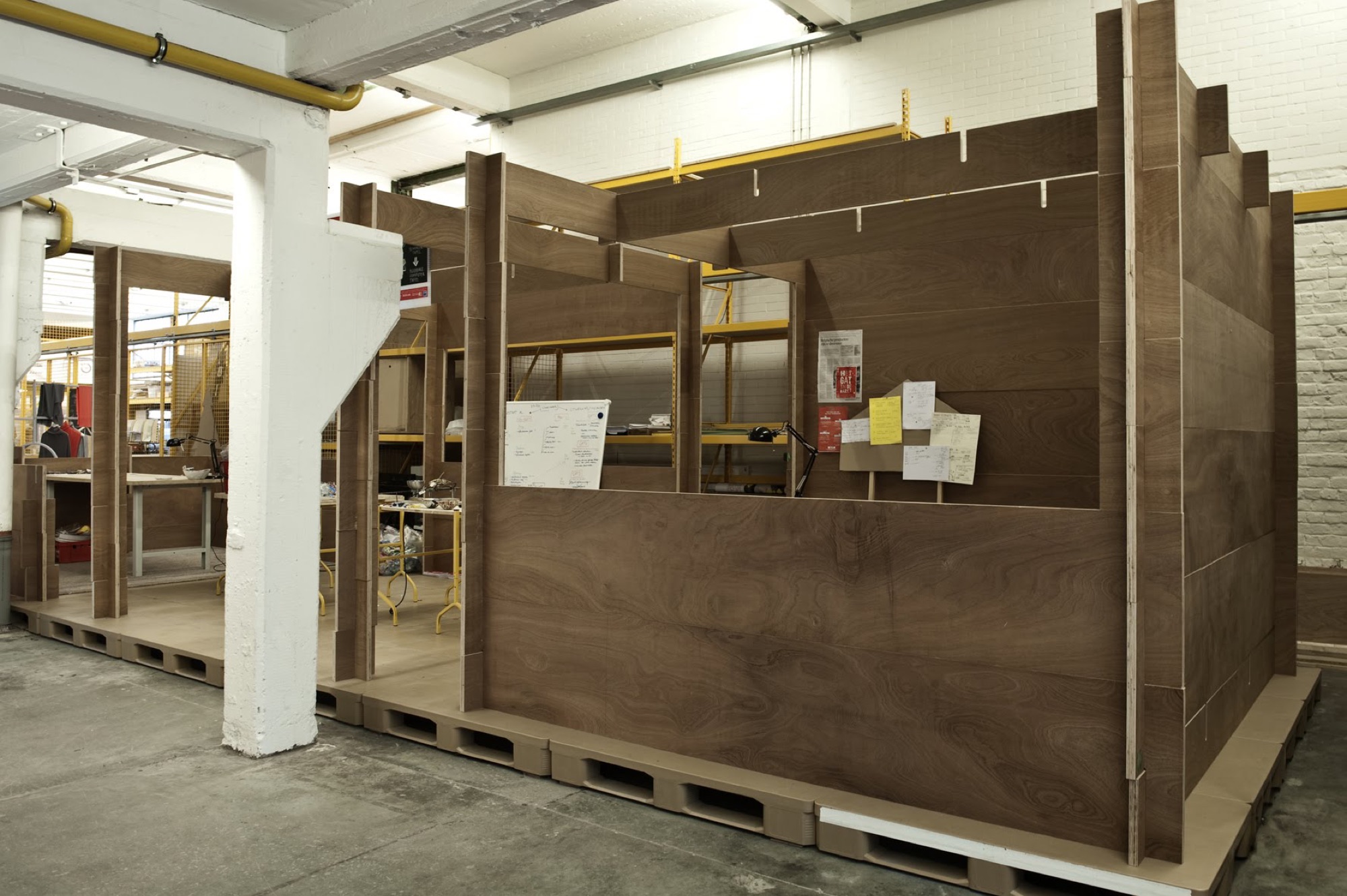 Houten modules op paletten in de fabriekshal van BUDA.lab, met werkplekken en aankondigingen tegen de witgeschilderde wanden