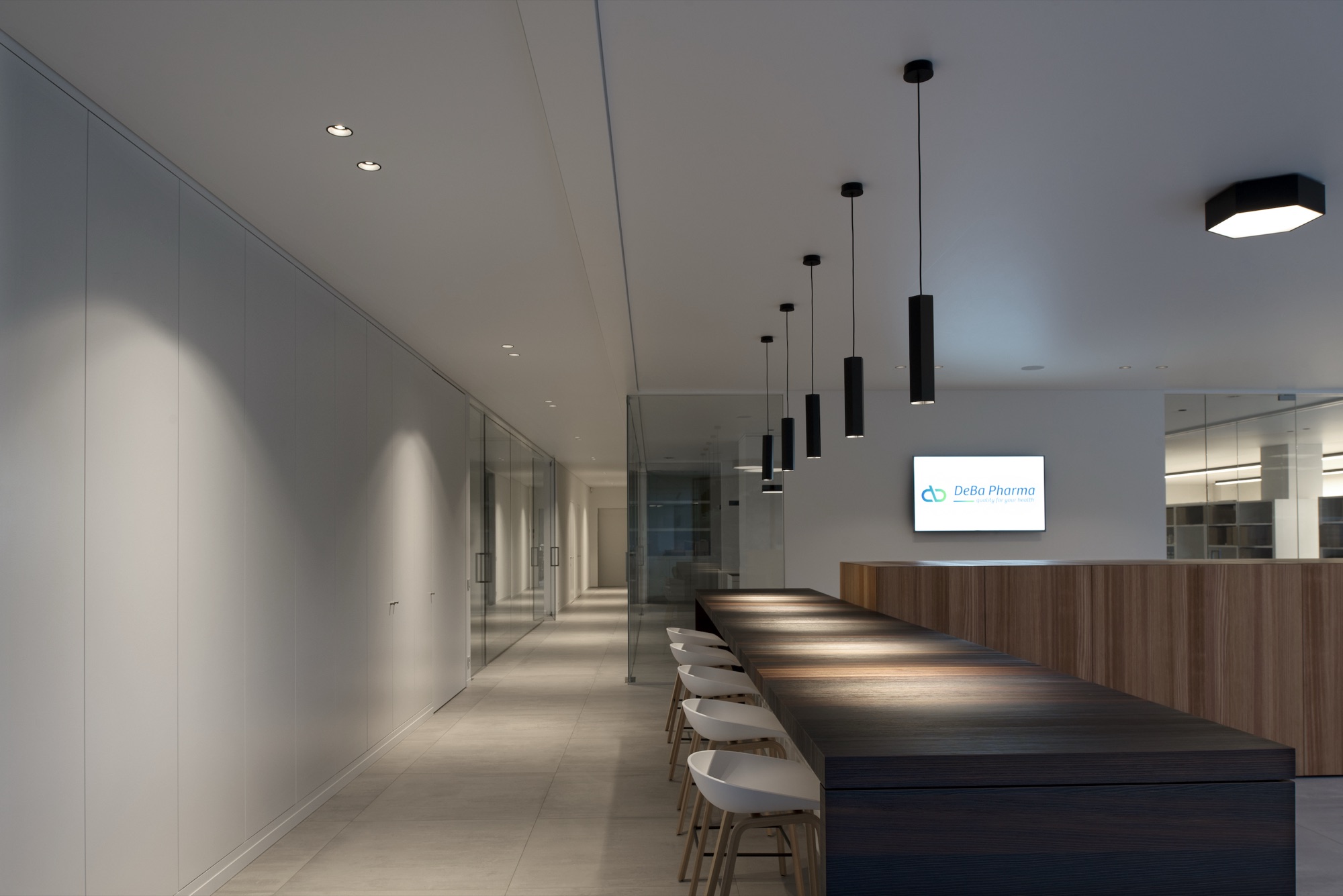 Lange gang bij DeBa Pharma met een doorlopende tafel met krukken, een rij zwarte hanglampen en een verlicht glaspaneel over de volle wandlengte