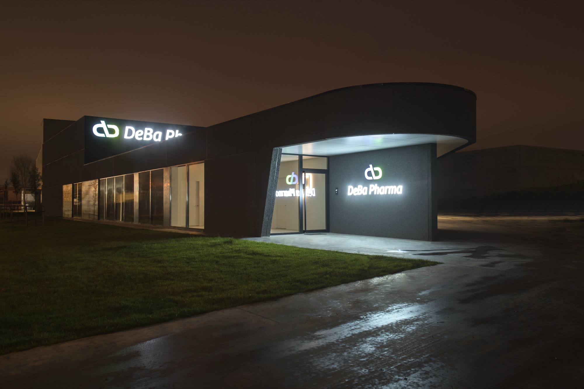 Gevel van DeBa Pharma bij avond: een donker volume met een gebogen luifel, verlichte belettering en een verlichte inkom boven een natte weg