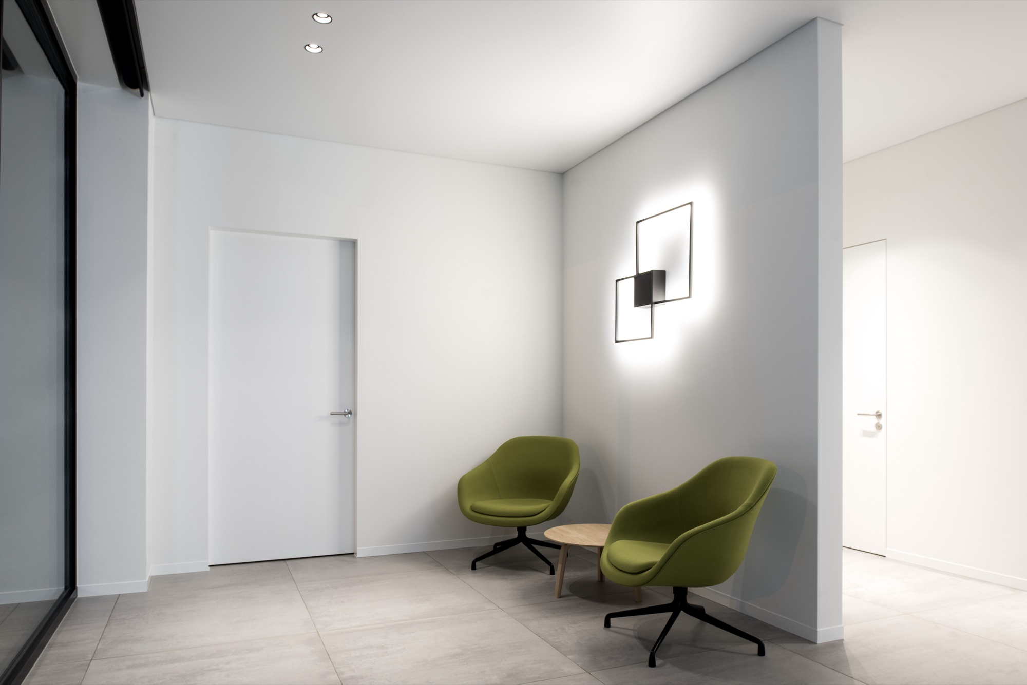 Wachthoek bij DeBa Pharma met twee groene fauteuils, een lage tafel, een verlicht wandobject en vlak ingewerkte witte deuren