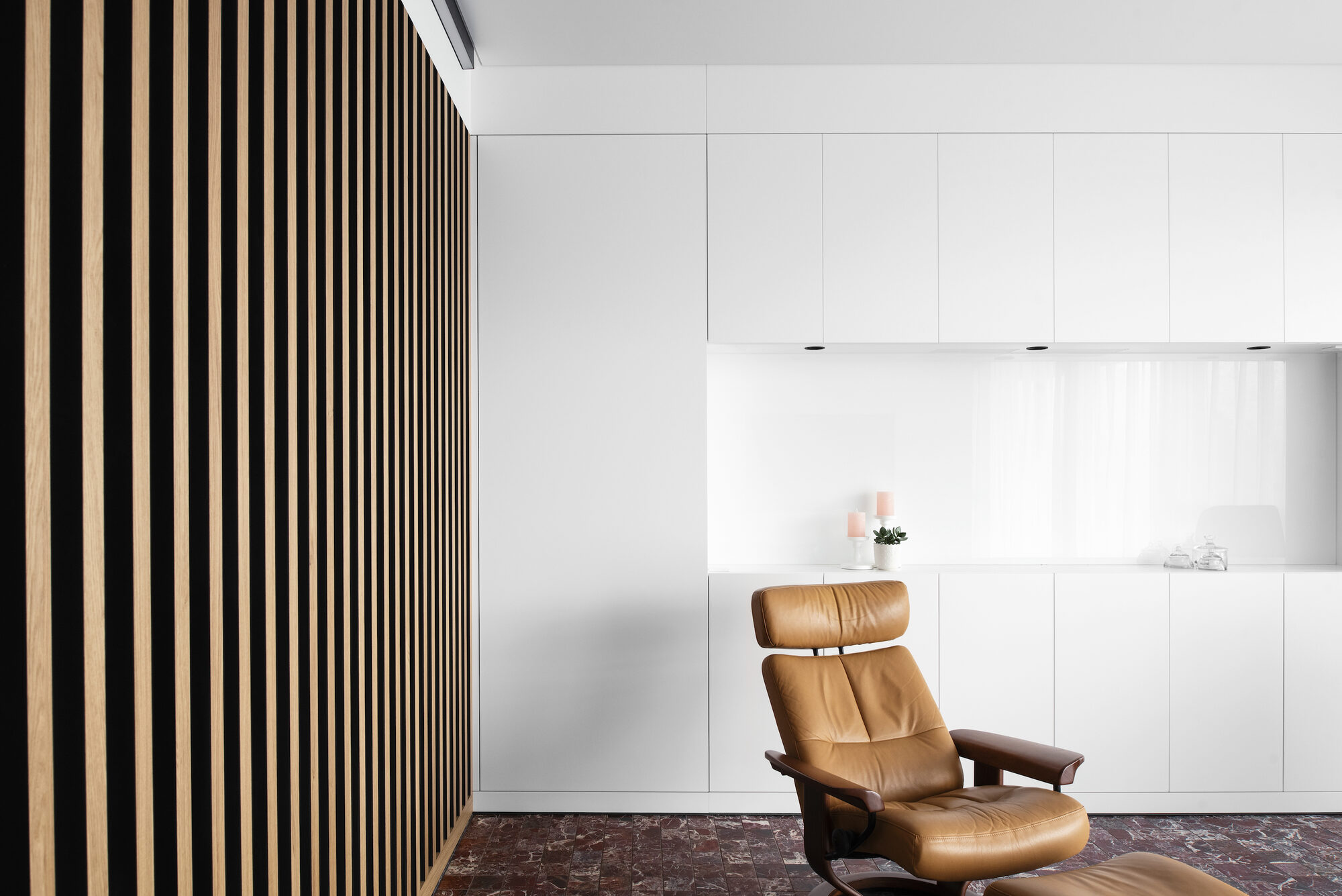 Interieur van Factory Appartement met een wand van verticale eiken latten links, een strakke witte keuken met doorlopende kastenwand rechts, een leren fauteuil en een vloer in rode natuursteentegels