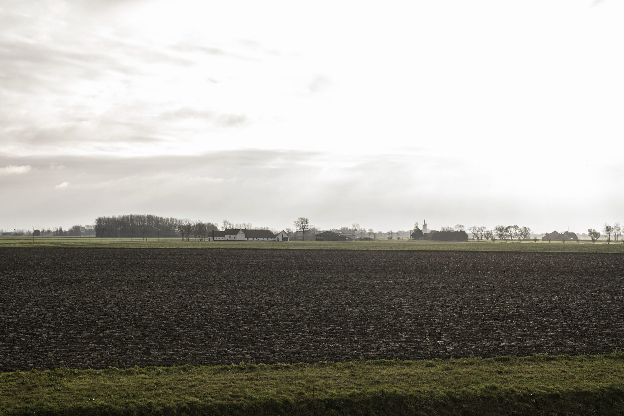 Open polderlandschap bij grijze lucht met een geploegd veld op de voorgrond en een verre bomenrij met hoevegebouwen aan de horizon