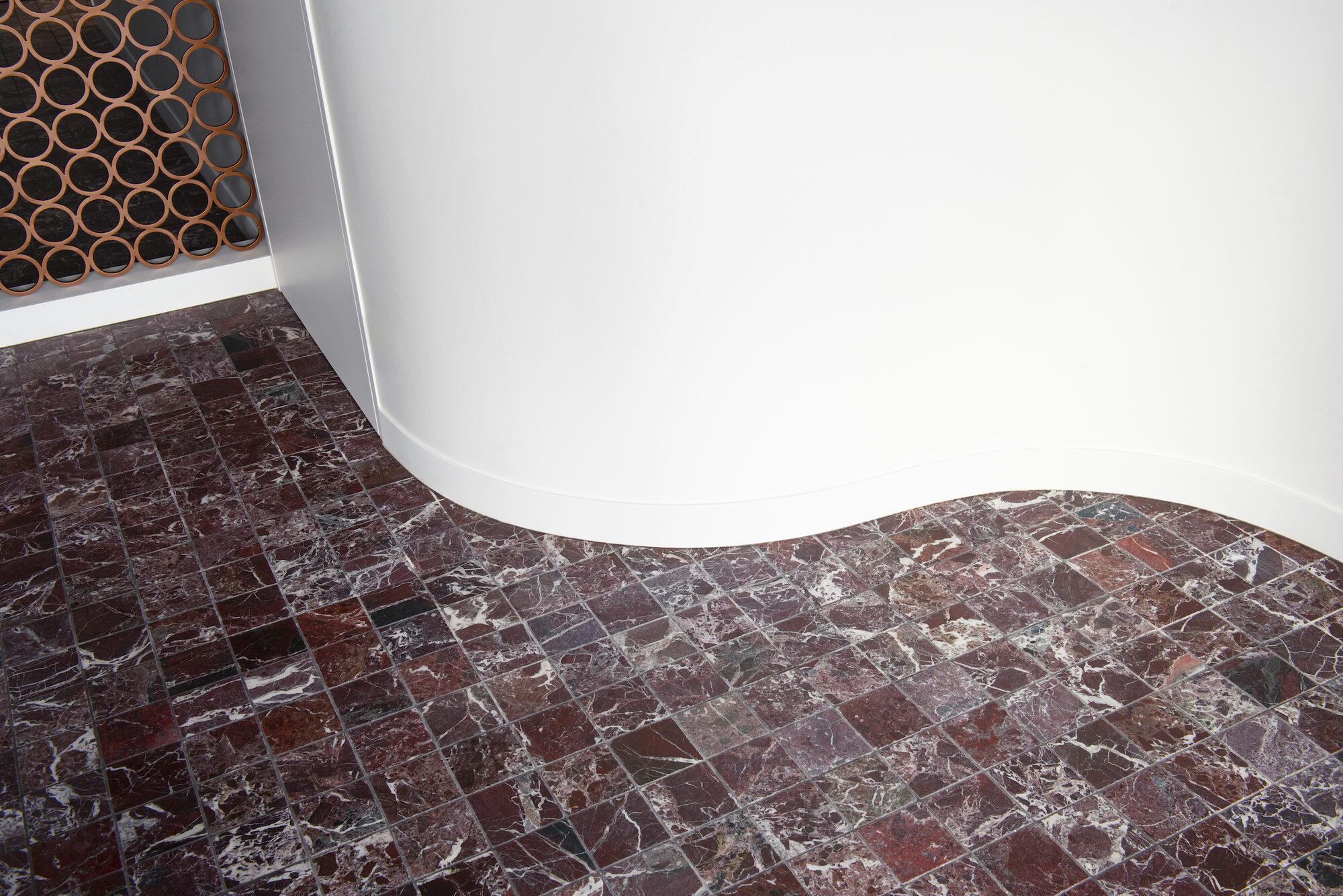 Detail in Factory Appartement: een vloer van rode natuursteentegels die in een boog langs een gebogen witte wand loopt, met links een koperkleurig rasterpaneel