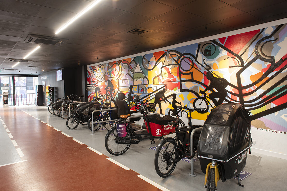 Stallingsruimte van Fietskot met bakfietsen aan beugels, een gemarkeerde rijstrook op de vloer en een grote kleurrijke muurschildering over de volle wand