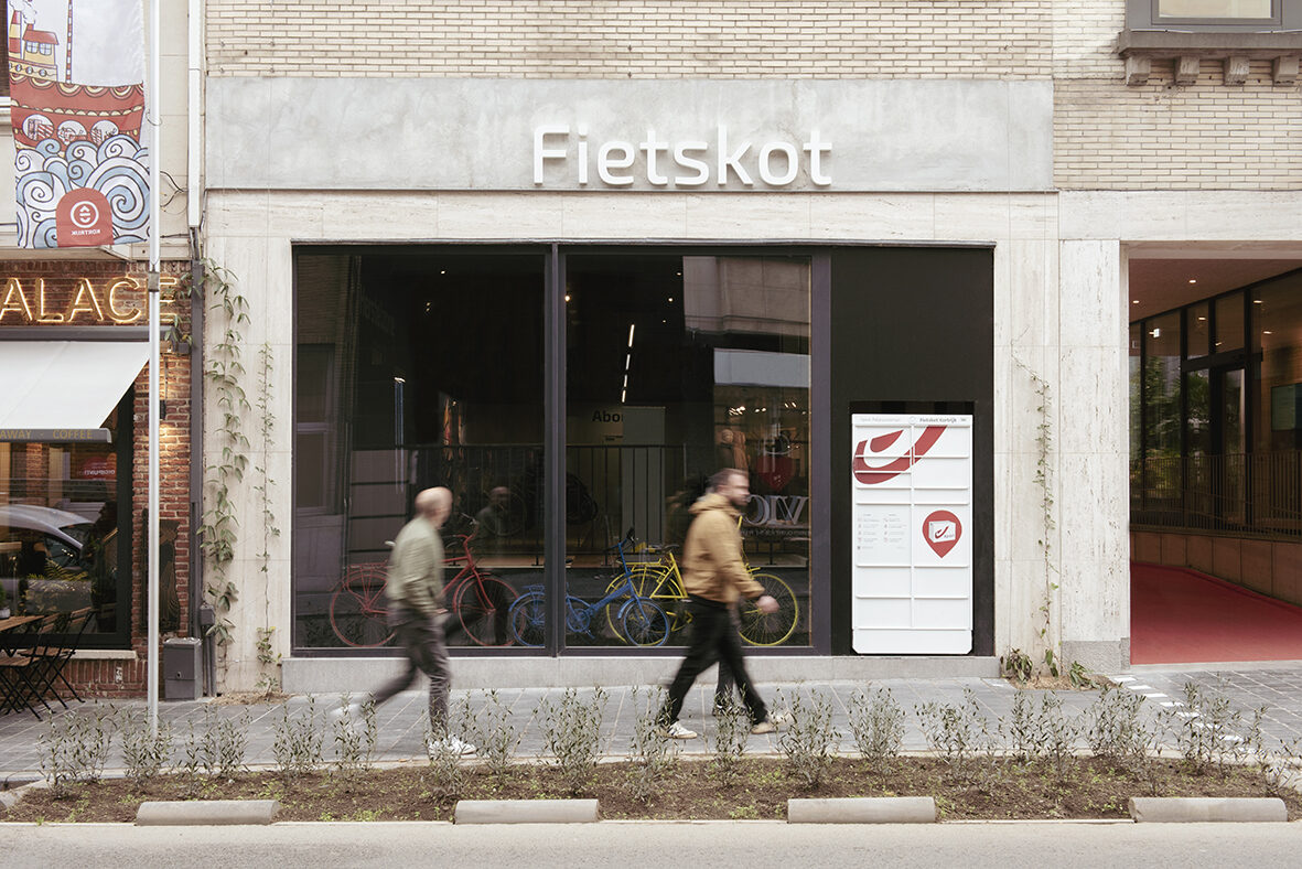 Straatgevel van Fietskot in Kortrijk met de naam in witte letters op een betonnen bovenlijst, een brede etalage met fietsen erachter, en twee voorbijgangers op het voetpad met een strook beplanting