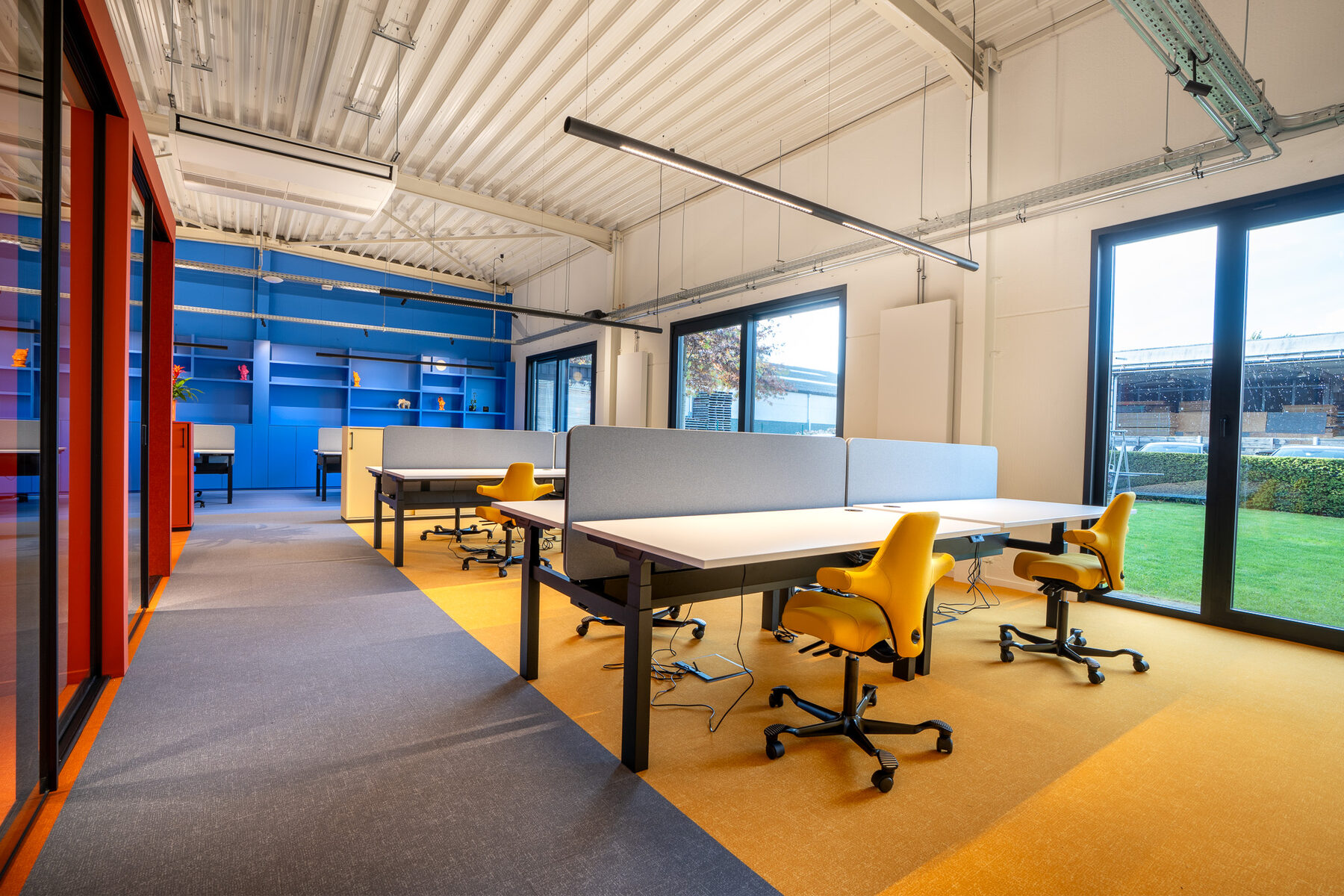 Landschapskantoor van GBL Agency met twee rijen bureaus met lage akoestische schermen, gele bureaustoelen, een gele tapijtzone onder de werkplekken en een blauwe maatkastwand achteraan