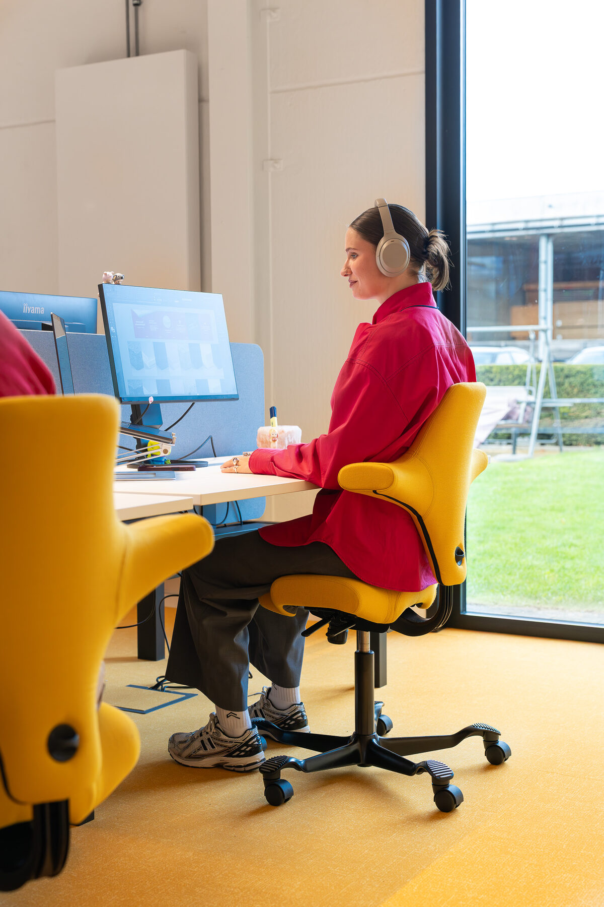 Medewerker met koptelefoon aan een bureau bij het raam in het kantoor van GBL Agency, met gele bureaustoelen op een gele vloerzone en zicht op het grasveld buiten