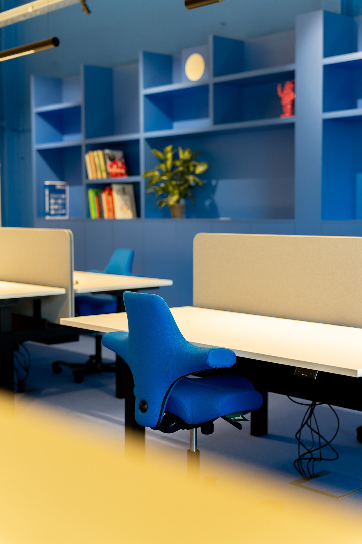 Detail van twee in hoogte verstelbare bureaus bij GBL Agency met beige akoestische scheidingsschermen, een blauwe bureaustoel en de blauwe vakkenkast op de achtergrond