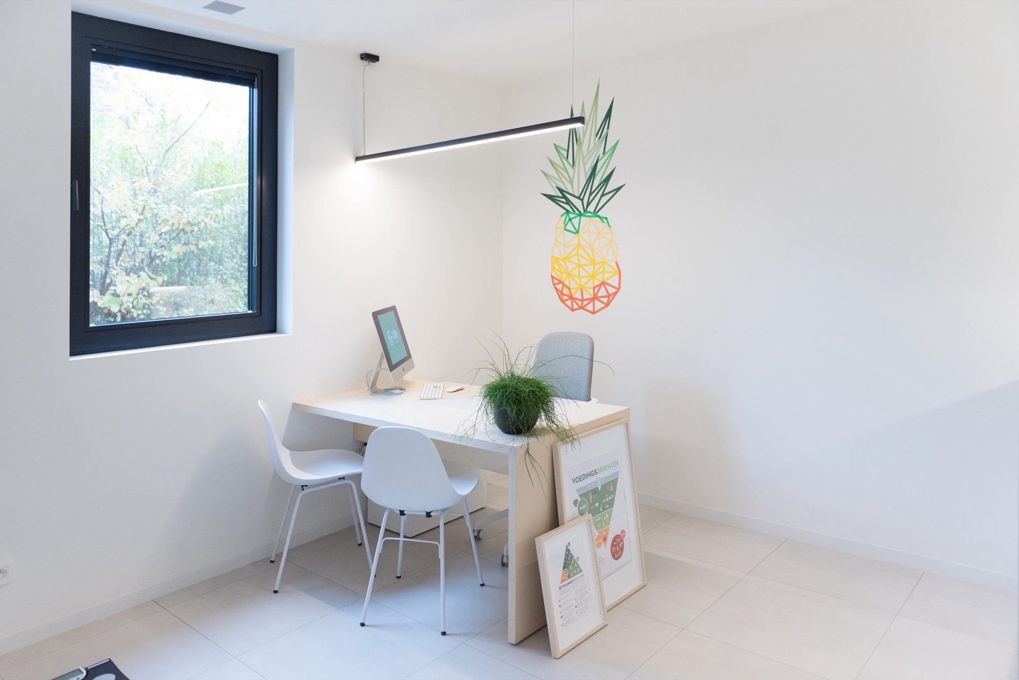 Kabinet in Groepspraktijk Tille met een licht bureau voor twee stoelen bij een raam op de tuin, een lijnarmatuur en een gekleurde muurillustratie