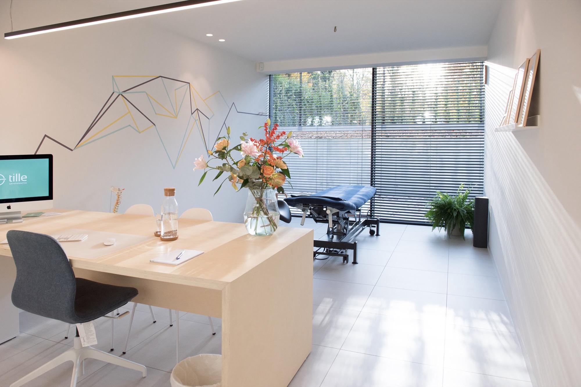 Kabinet in Groepspraktijk Tille met een houten bureau vooraan en een donkere behandeltafel achteraan bij een raam met lamellen