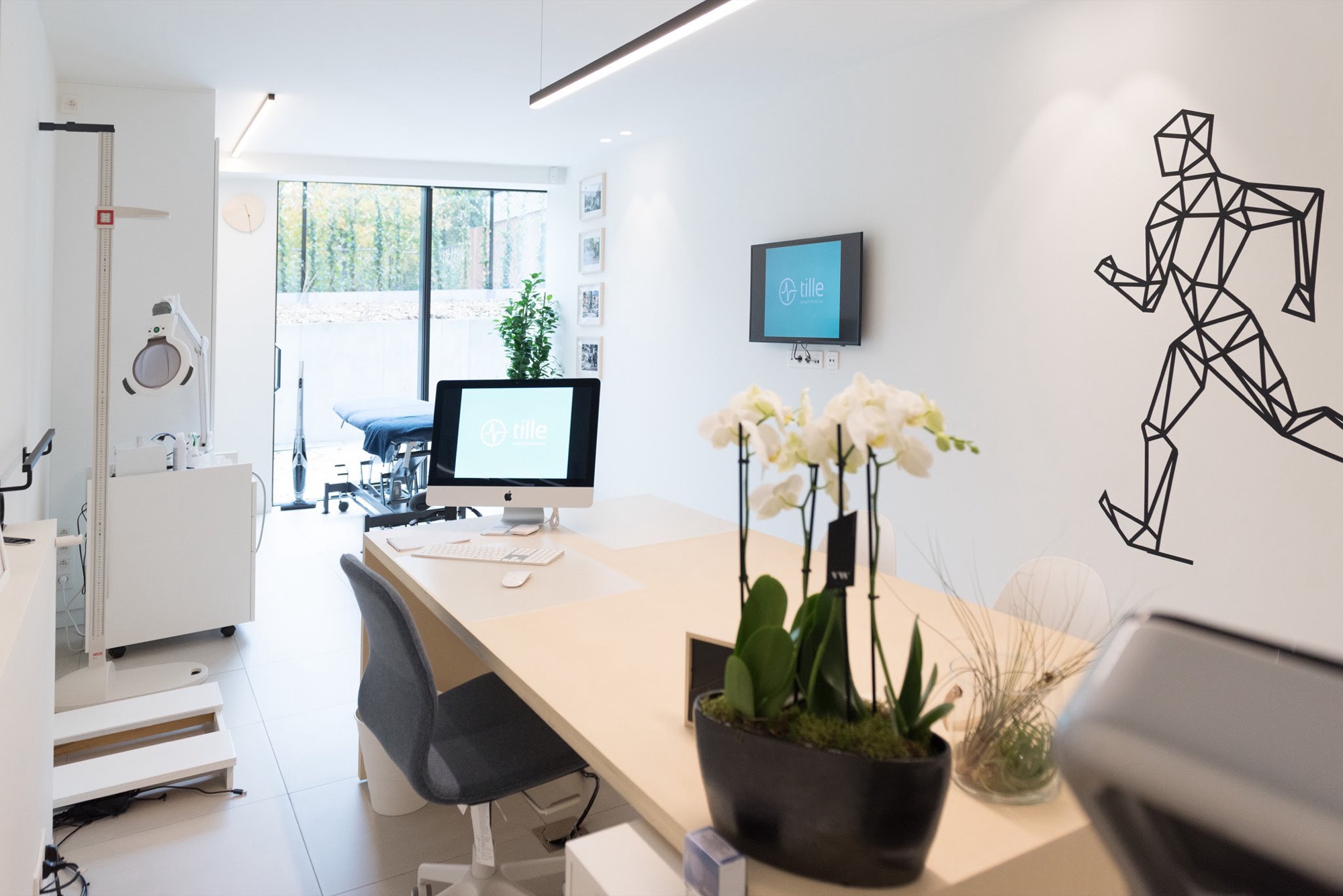 Kabinet in Groepspraktijk Tille met een lange werktafel, een wandscherm, medische apparatuur langs de wand en daglicht via een raam over de volle hoogte