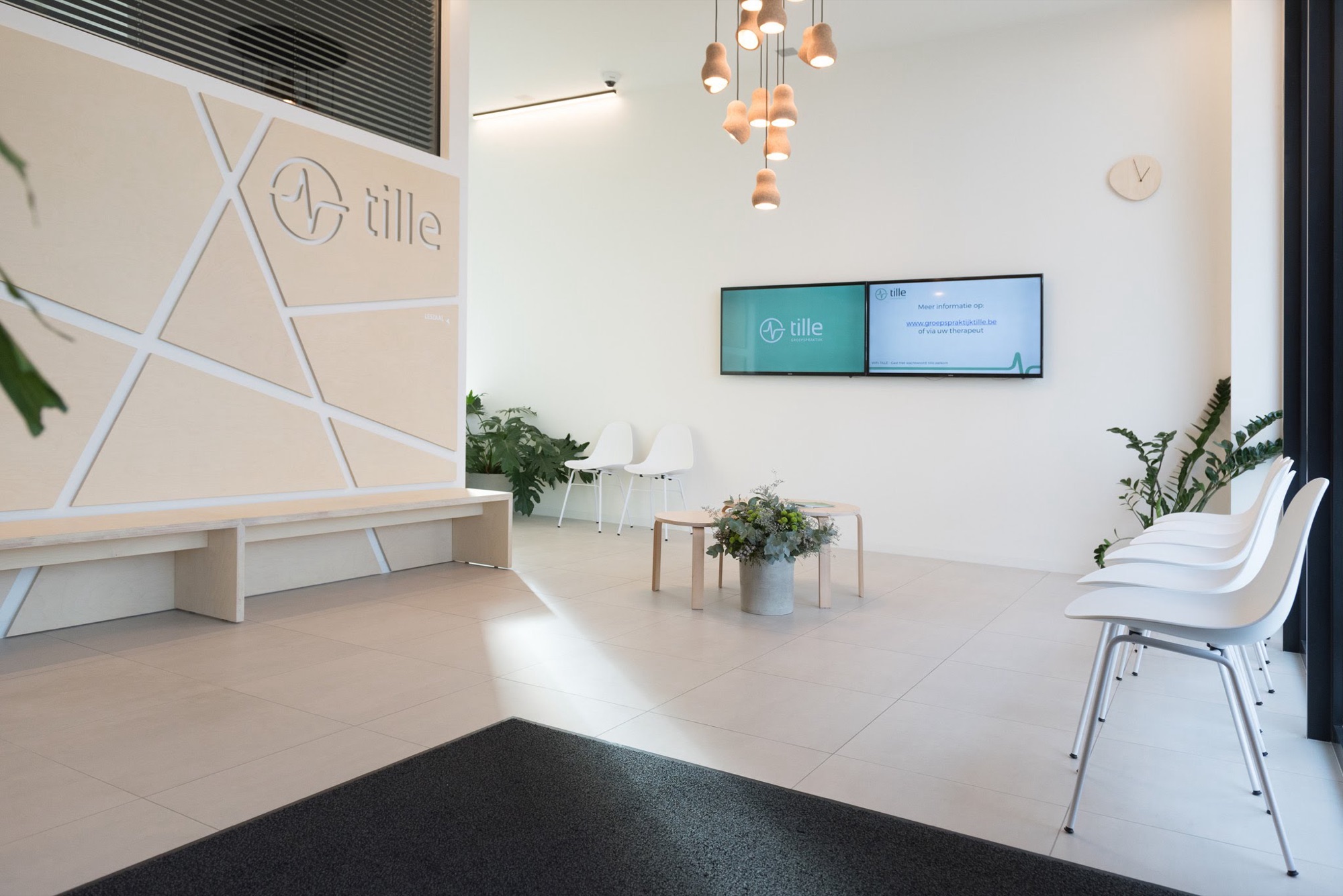 Onthaal van Groepspraktijk Tille met het logo op een lichte wand met ingefreesde lijnen, een ingewerkte zitbank, twee wandschermen en een rij witte wachtstoelen