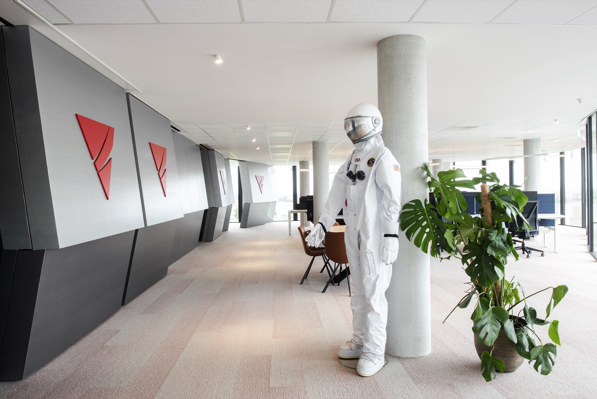 Gang bij Ingenix met afwisselend donkere en lichte wandpanelen met het rode logo, een betonnen kolom en een astronautenpak als decorstuk
