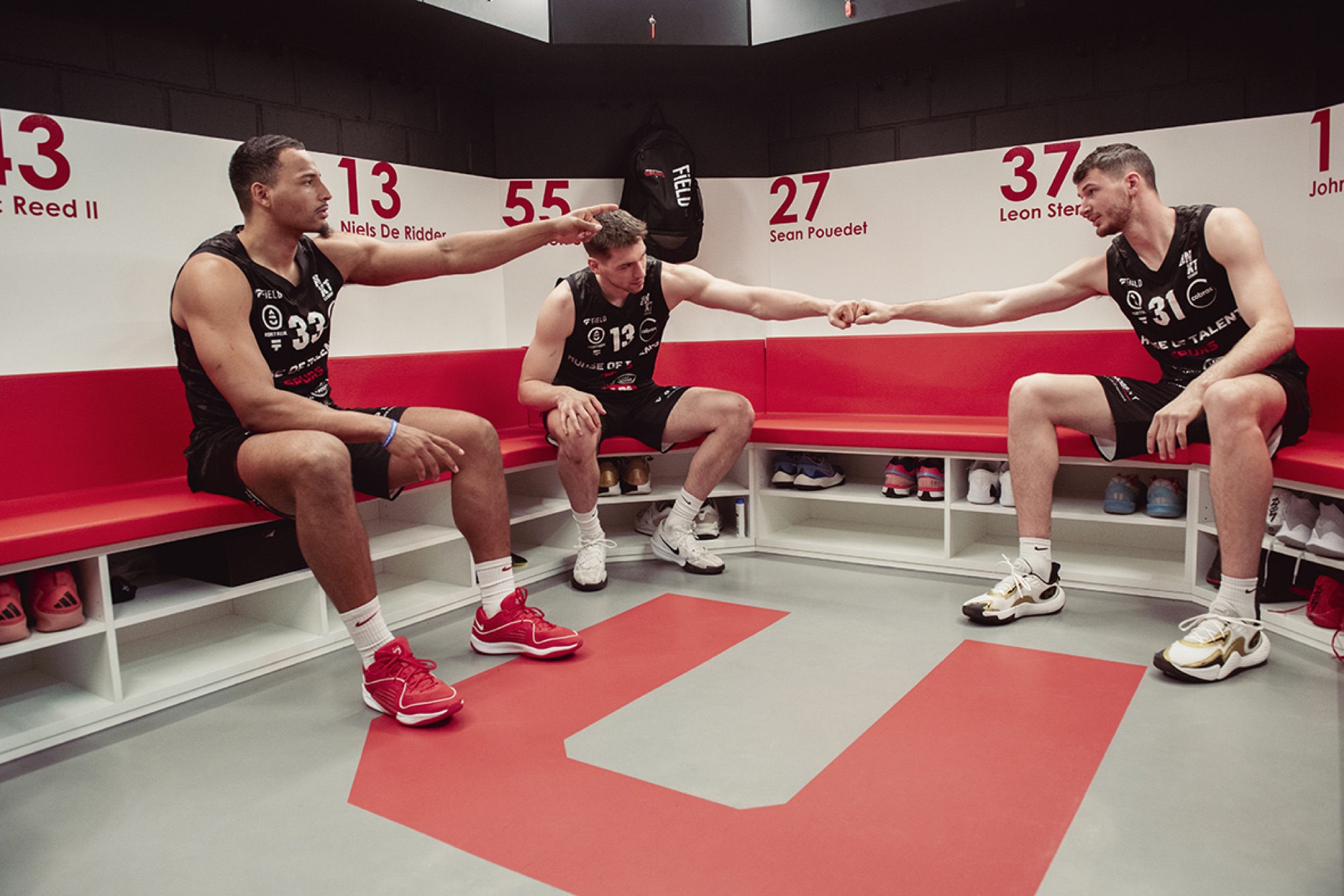 Drie spelers van de Kortrijk Spurs op de rode zitbank in de kleedkamer, met het clublogo in de vloer
