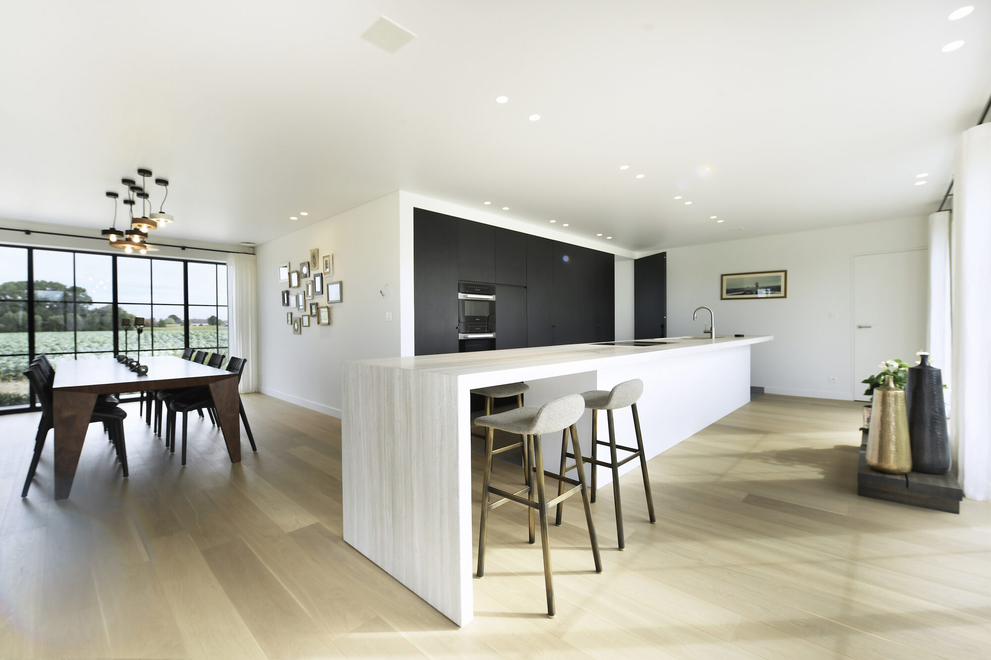 Open leefkeuken van Modern Farmhouse met een lang keukeneiland met steenkleurige zijwang, twee krukken, een zwarte kastenwand met ingebouwde toestellen en links een houten eettafel bij het raam op het veld