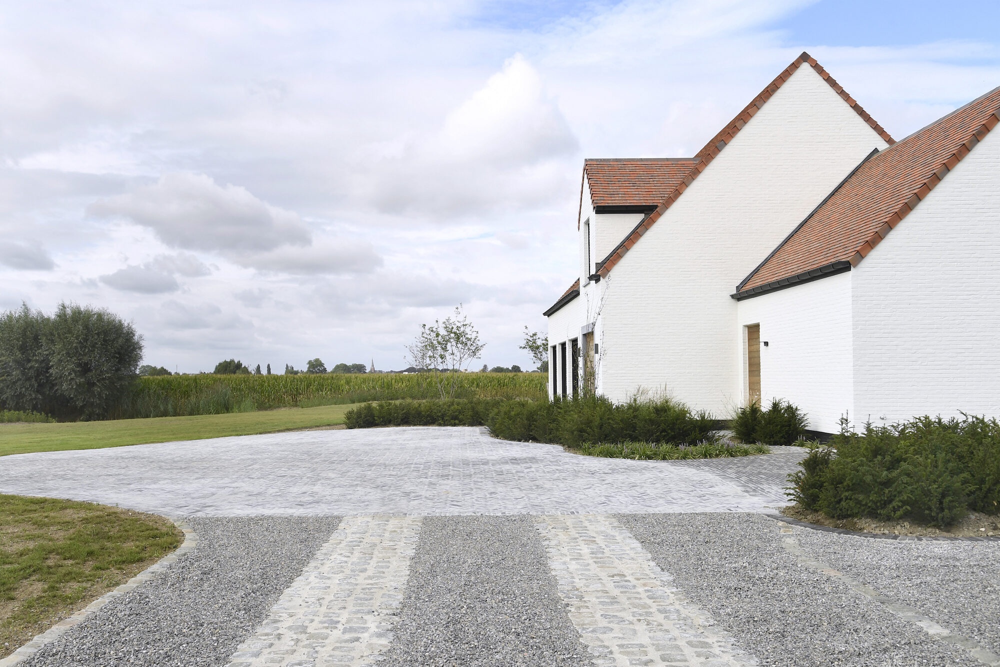 Modern Farmhouse vanaf de oprit: twee witte volumes met puntgevels en rode pannendaken, een oprit in grind met kasseistroken, lage groenaanplant tegen de gevel en open velden ernaast