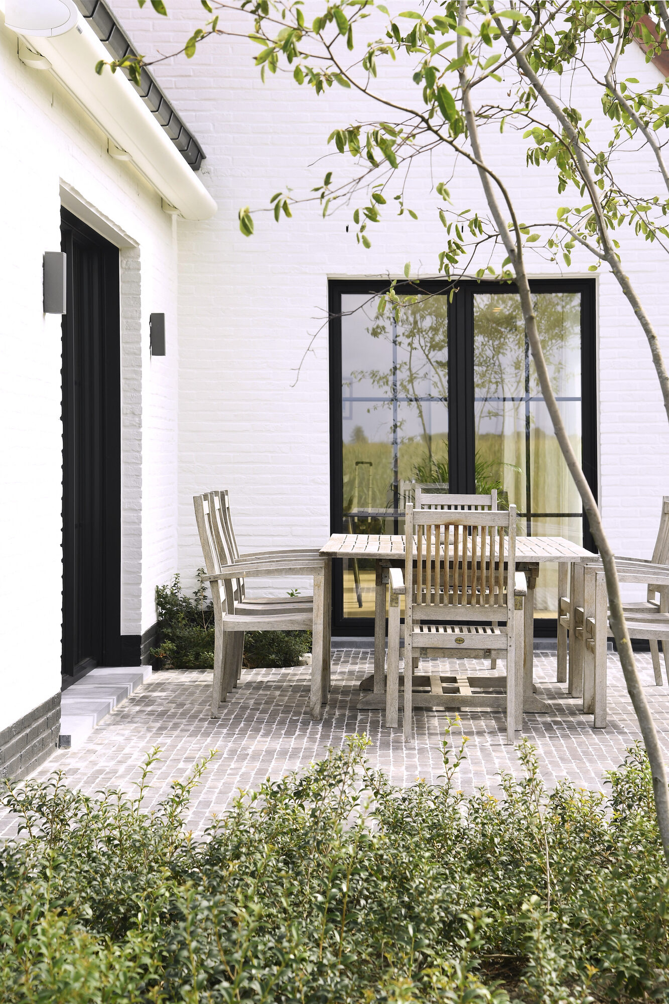 Beschut terras van Modern Farmhouse met een houten tuintafel en stoelen op kleinschalige kasseien, tegen een witte gevel met zwarte schuiframen en een jonge boom ervoor