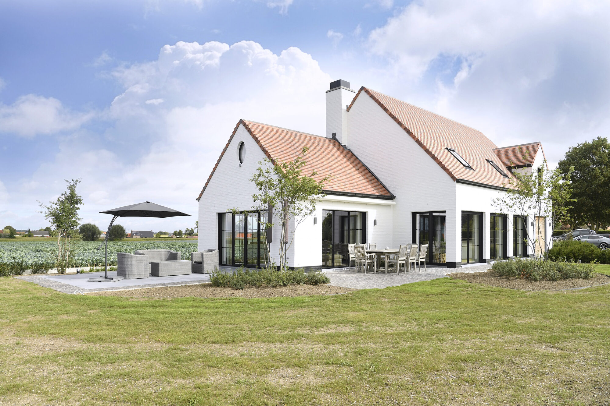 Tuingevel van Modern Farmhouse: een witte nieuwbouwhoeve met rood pannendak, doorlopende zwarte raampartijen, een terras met eettafel en parasol, en een gazon dat overgaat in het omliggende veld