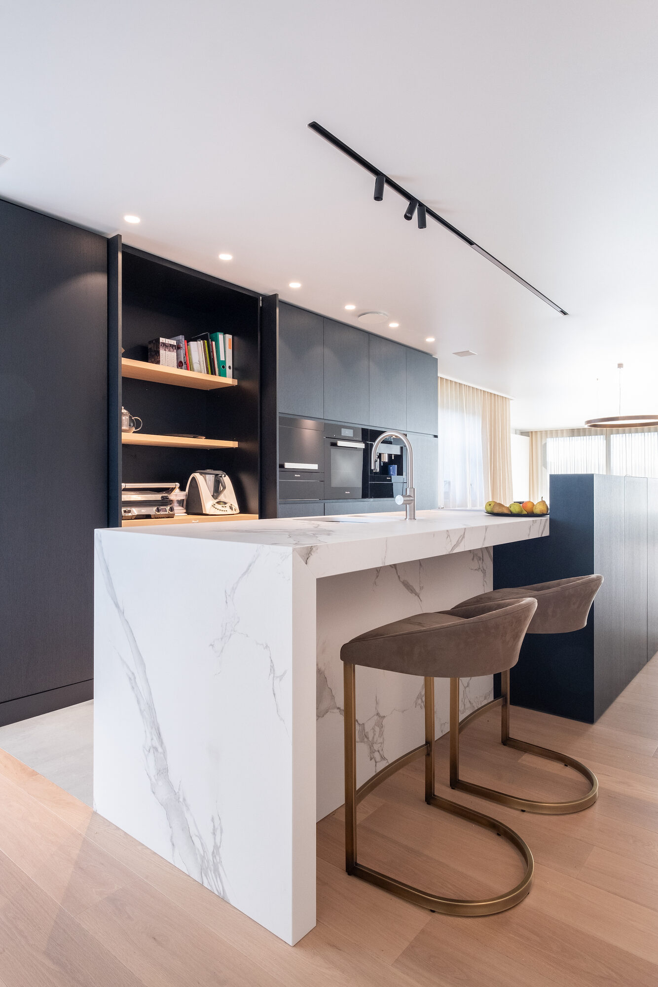 Keuken van New Winehouse met een keukeneiland in wit marmerdecor, twee velours krukken op messingkleurige poten, een donkere kastenwand met open eiken vakken en een zwarte lichtrail aan het plafond