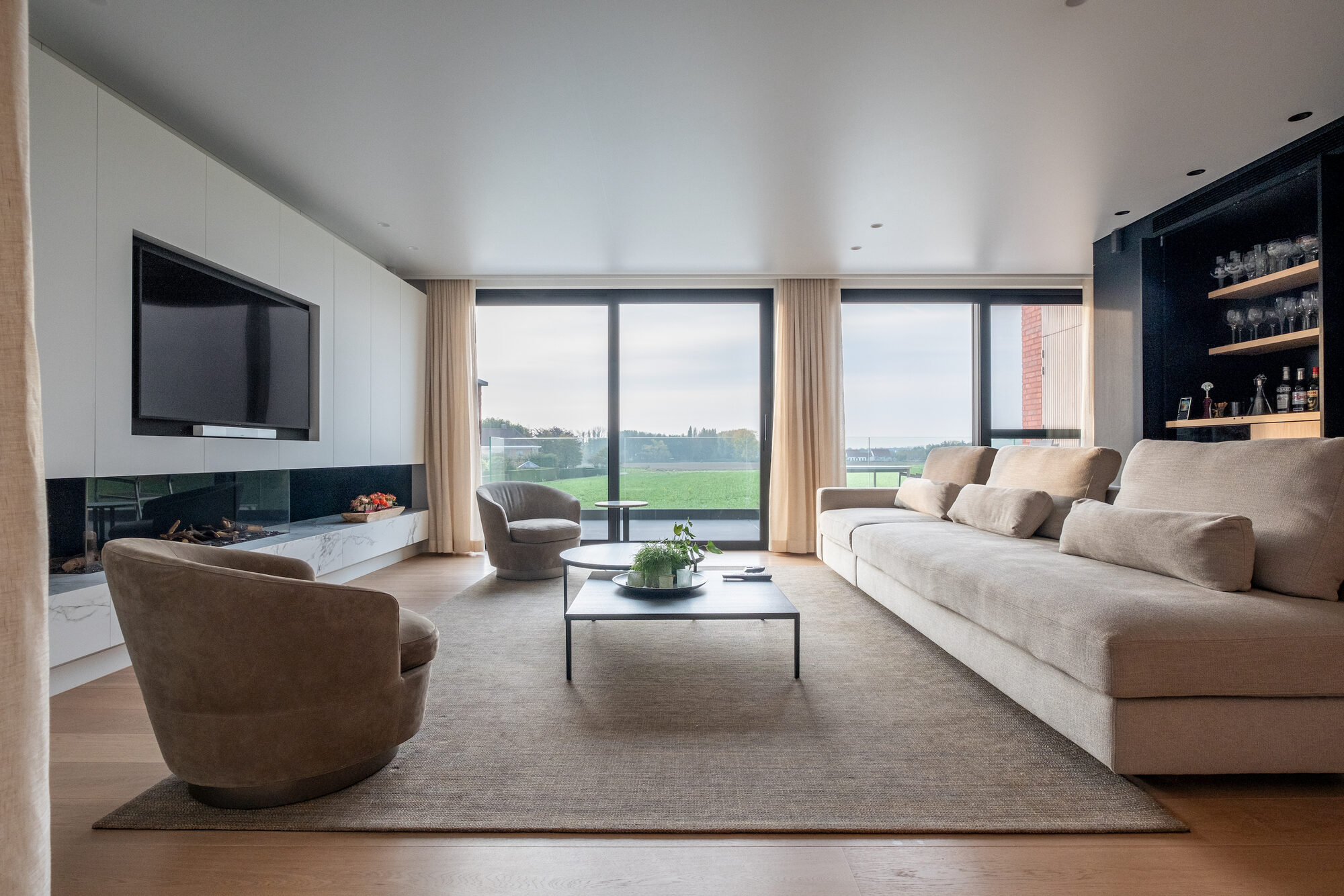 Leefruimte van New Winehouse met een lange lichte zetel, twee ronde fauteuils, een ingewerkte haard onder een tv in een witte kastenwand en schuiframen met zicht op de velden