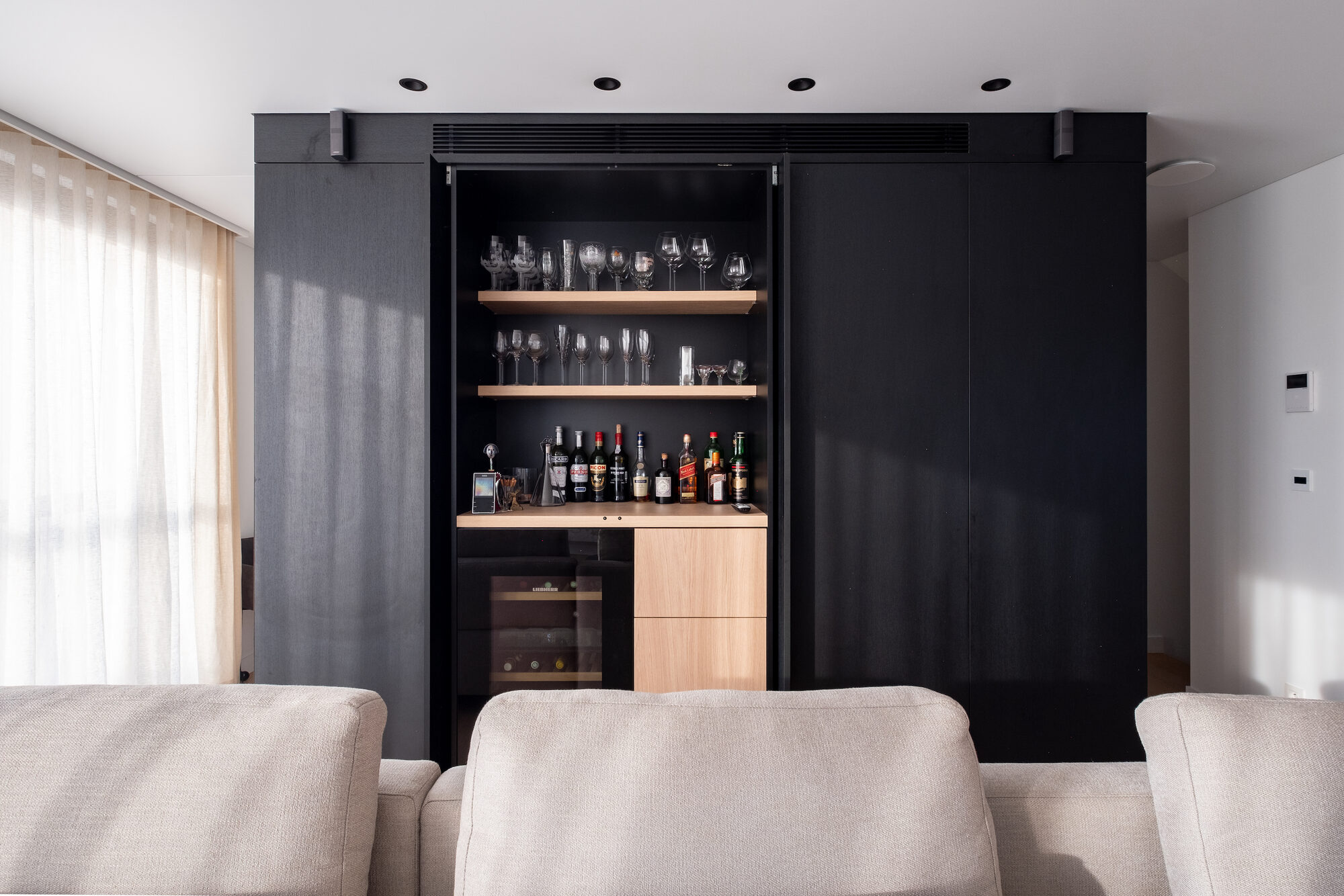Zwarte wandkast in New Winehouse met schuifpanelen die openschuiven op een barnis met eiken legplanken vol wijnglazen, flessen op een eiken blad en een ingebouwde wijnkoelkast eronder