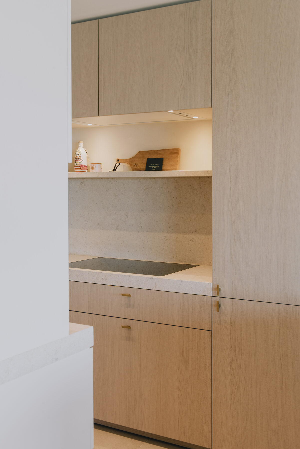 Keukenwand in het appartement in Nieuwpoort in licht eik met een verlichte nis, een stenen achterwand en een kookplaat verwerkt in het lichte werkblad