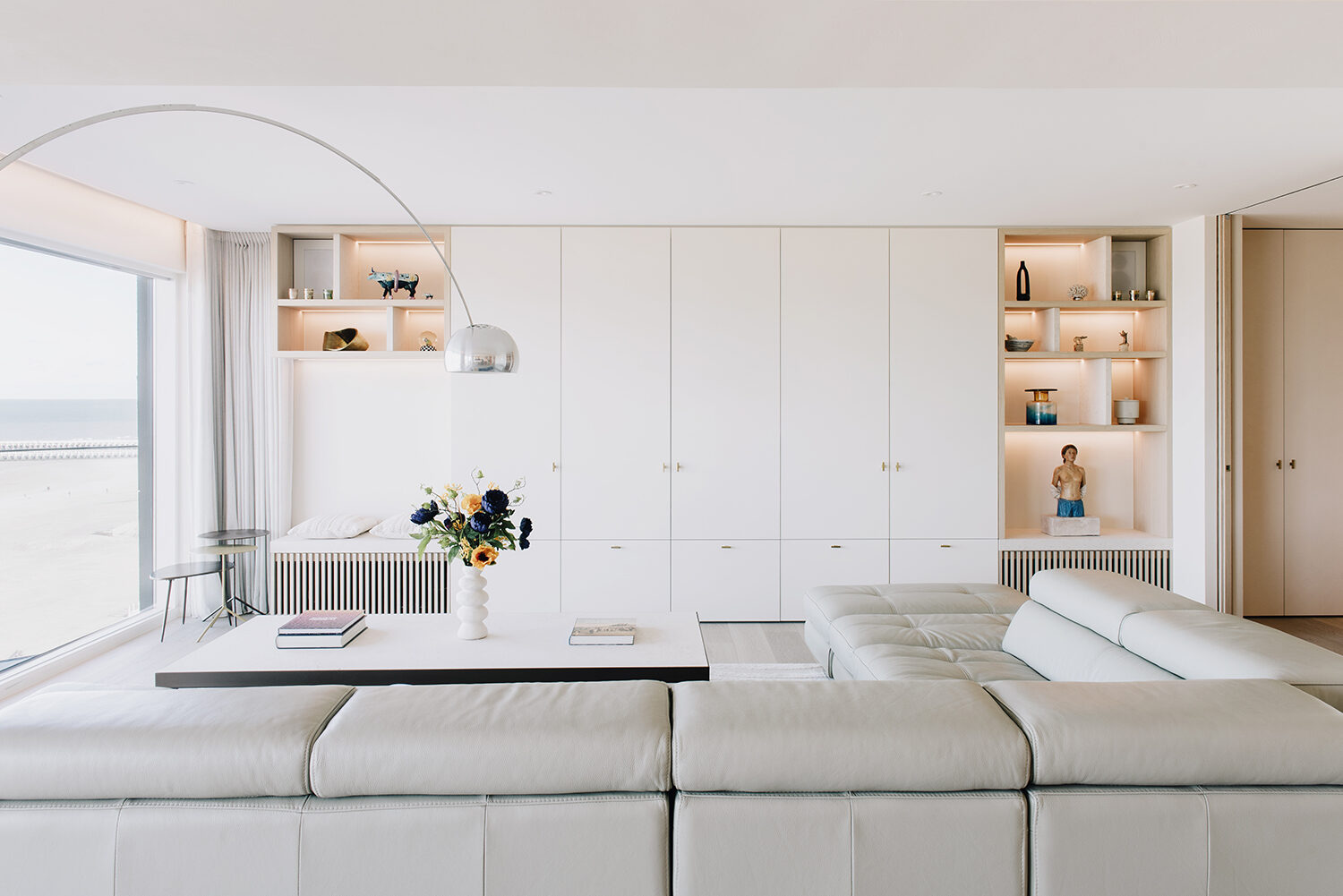 Leefruimte in het appartement in Nieuwpoort met een lichte modulaire zithoek en een witte kastenwand met verlichte nissen